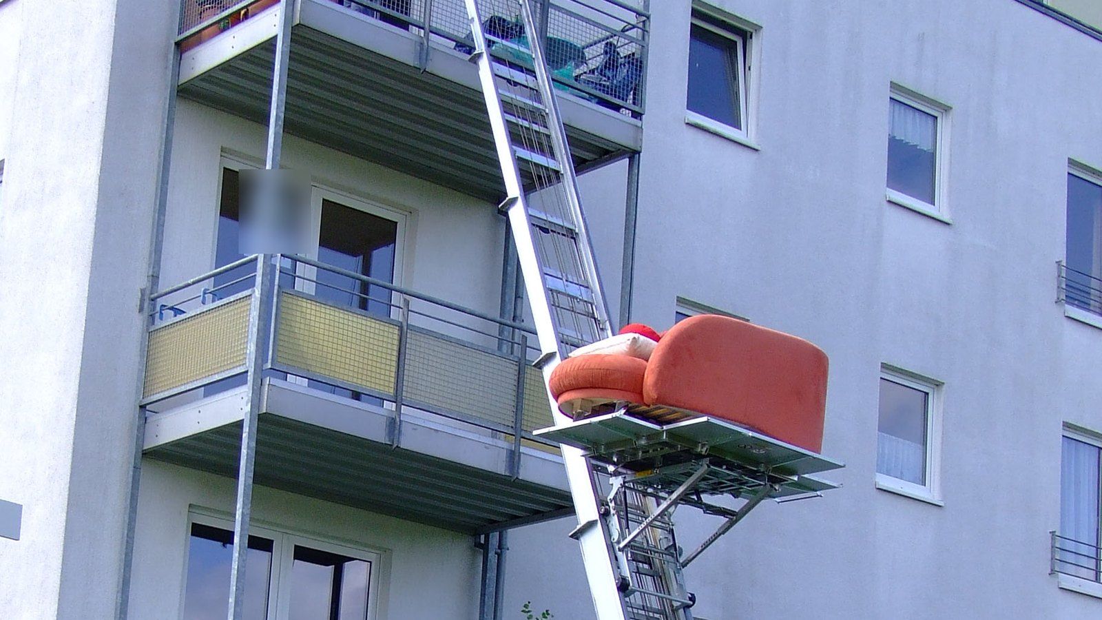 Ein Möbellift an einem Wohnhaus bringt ein Sofa auf der Plattform zum Balkon.